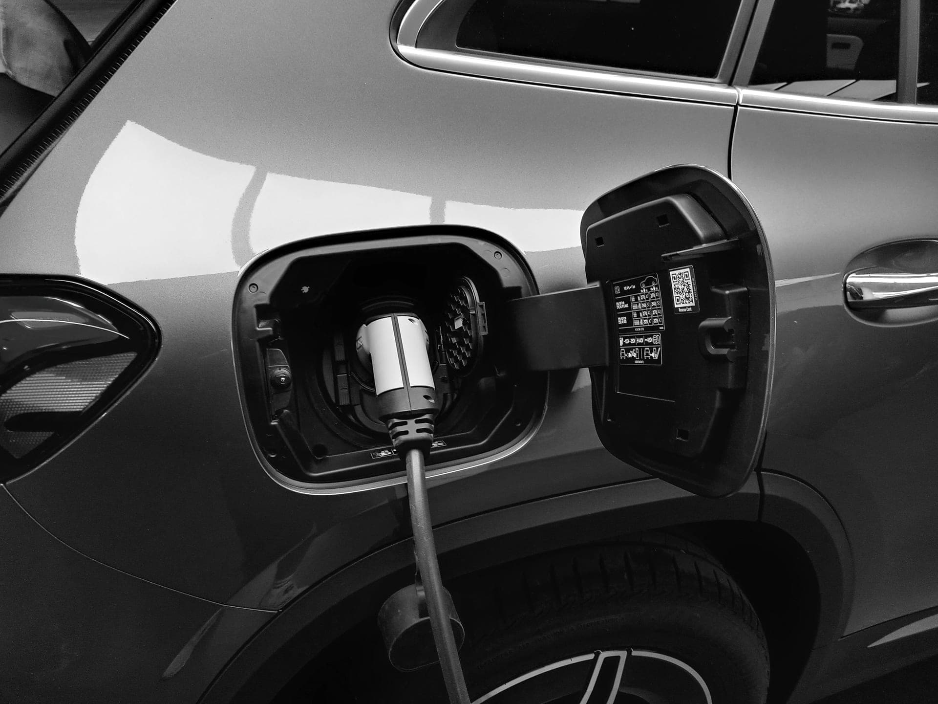 A charging connector plugged into an electric car's charge port, its voltage-spec label visible on the port door